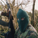 IRAN Arming Irish Militants — Nobody Saw This Coming… Individual in camouflage holding a rifle, wearing a green balaclava
