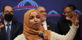 Ilhan Omar speaking passionately at a podium with a group of supporters behind her