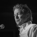 Rand Paul’s Son Caught in HORRIFYING Outburst… A man with curly hair speaking into a microphone in black and white