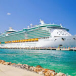 Cruise Line Downplays DISASTER—115 Fall Ill… Large cruise ship docked near turquoise water and foliage.