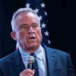 Washington Knifes RFK Jr.’s Vaccine Shake-Up Man in suit holding a microphone, American flag background.