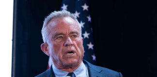 Man in suit holding a microphone, American flag background.
