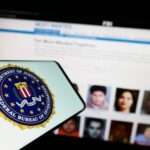 Medicare Heist MASTERMIND FINALLY Nabbed Abroad FBI logo displayed on a smartphone with a website showing wanted individuals in the background