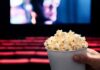 A hand holding a cup of popcorn in a movie theater with a blurred screen in the background