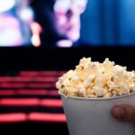 Hollywood’s $10B Mirage Exposed A hand holding a cup of popcorn in a movie theater with a blurred screen in the background
