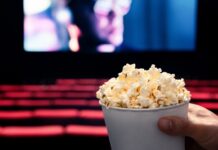 Hollywood’s $10B Mirage Exposed A hand holding a cup of popcorn in a movie theater with a blurred screen in the background