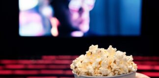 A hand holding a cup of popcorn in a movie theater with a blurred screen in the background