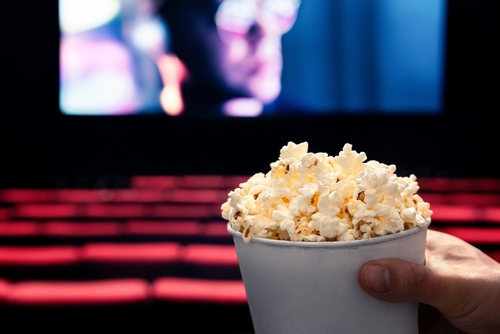 Movies,And,Popcorn.,Man,Holding,Pop,Corn,Box,At,Cinema. A hand holding a cup of popcorn in a movie theater with a blurred screen in the background