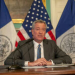 Shocking U-Turn: De Blasio Praises Trump Tactic Man speaks at a press conference with flags behind.