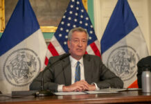 Shocking U-Turn: De Blasio Praises Trump Tactic Man speaks at a press conference with flags behind.