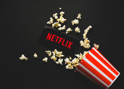shutterstock_1828714433 Spilled popcorn, phone displaying Netflix logo, striped popcorn container.