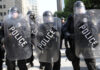 Police in riot gear holding shields in a formation.