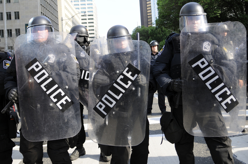 Police in riot gear holding shields in a formation.