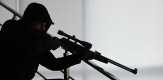 Person in dark clothing aiming a rifle indoors.